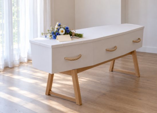 Plain white cardboard coffin with rope handles on a wooden bier, with a small posy of cornflowers and daisies on the lid