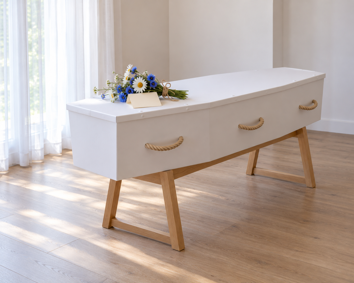 Plain white cardboard coffin with rope handles on a wooden bier, with a small posy of cornflowers and daisies on the lid